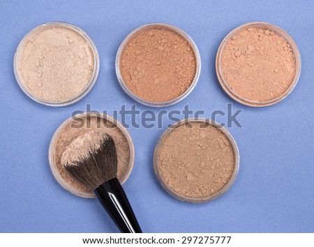 Close-up of makeup brush and jars filled with loose cosmetic powder different shades on blue background. Top view