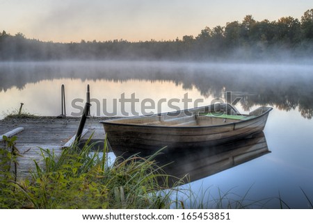 A dirty boat is parked by a wooden pier on a calm lake. It is dawn and soon the sun will be rising.