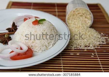 Cooked white rice with spices star anise ,onion rings and tomatoes served on a white plate on a bamboo mat and  raw rice in the background .sticky rice and scattered basmati or jasmine rice. garnished