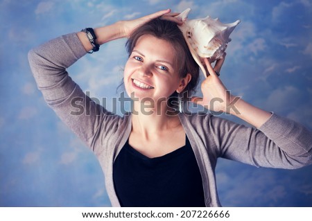 attractive blue-eyed girl head cocked smiling and looking at the camera holding a conch shell on her head as a hat on a blue background