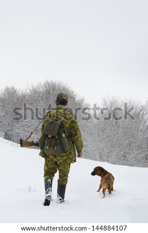 Male hunter in camouflage clothes walking on the snow field with hunting rifle during a hunt, dog follows him, foggy weather