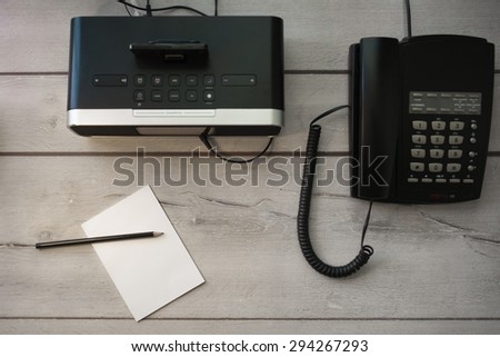 Equipment in a hotel room: Radio alarm clock, land line telephone, notepad and pencil, on aged wooden table.