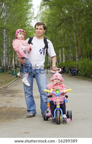 Father walking with their daughters along the way in a park: elder girl  is on his hands, younger ride on a tricycle