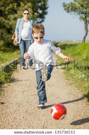 Happy cute son playing with soccer ball in the park, and his pregnancy mother looking in the background