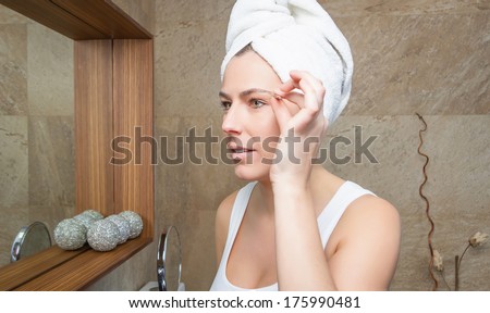 Beautiful young woman plucking her eyebrows with tweezers in front of mirror on the bathroom