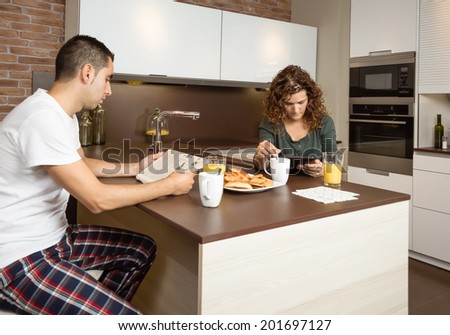 Serious young couple reading news in a digital tablet and newspaper while having breakfast on the home kitchen