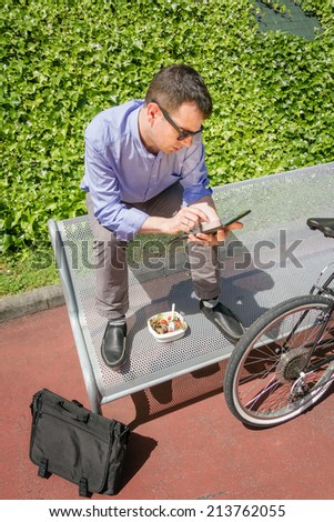 Young business man working with electronic tablet while having a lunch break, sitting on a bench outdoors