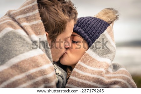Closeup of young beautiful couple kissing under blanket in a cold day with sea and dark cloudy sky on the background