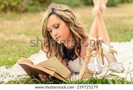 Portrait of romantic young woman with her shoes in the hand reading a book lying down over the grass. Selective focus on the shoes.