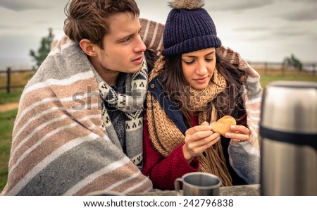 Portrait of young beautiful couple under blanket eating muffin in a cold day with sea and dark cloudy sky on the background