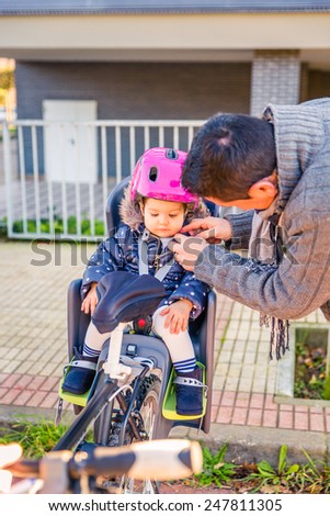Portrait of father closing bicycle security helmet to her little daughter sitting in bike seat. Safe and child protection concept