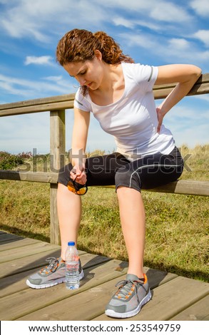 Athletic young woman in sportswear sitting touching her lower back muscles by painful injury, over a nature background. Sport injuries concept.