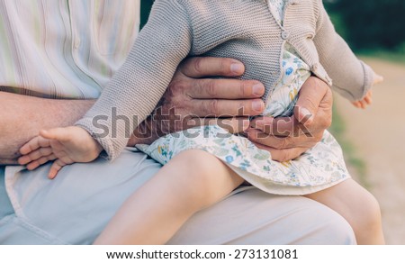 Closeup of baby girl sitting over legs of senior man outdoors. Two different generations concept.
