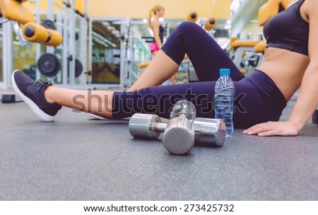 Closeup of sporty woman resting sitting on the floor of fitness center and female friend doing exercises with dumbbells in the background. Selective focus on a dumbbells.
