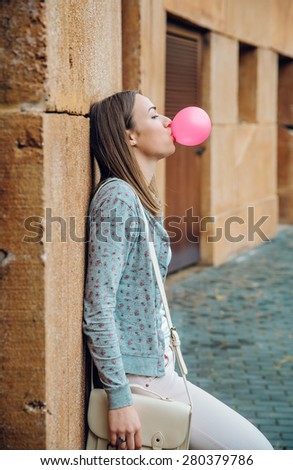 Portrait of beautiful young brunette teenage girl blowing pink bubble gum resting over a stone wall