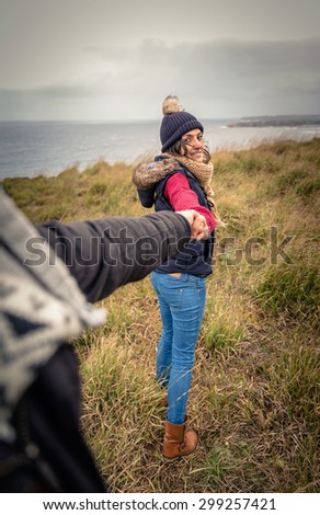 Back view of young beautiful woman with hat and scarf holding hand of man and leading by a meadow. Follow me to concept.