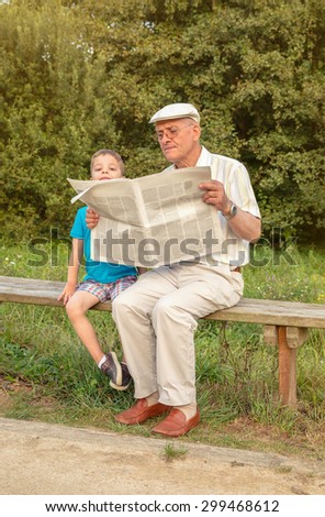 Senior man reading and bored cute child looking over the newspaper sitting on park bench. Two different generations concept.