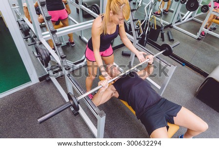 Woman personal trainer helping to muscle man for a correct bench press training with barbell on fitness center
