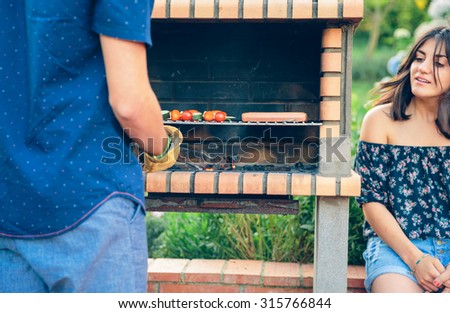 Back view of young man cooking sausages and vegetable skewers in a brick barbecue while woman looking in a outdoors summer party