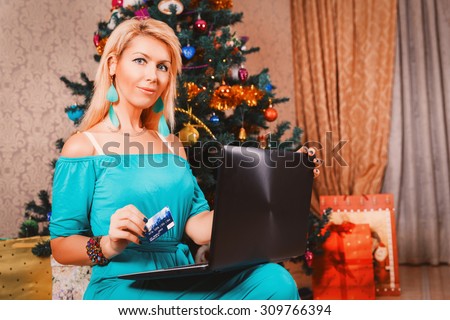 Pretty woman with laptop and credit card buying presents near Christmas tree