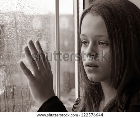 Stunning photo of a young girl gazing through a rainy window with her hand raised in sadness