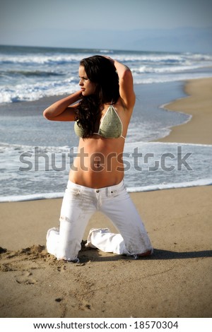 Sexy brunette kneeling on the sand in dirty jeans and bikini top
