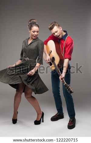 cheerful young caucasian guitar player and dancing girl in vintage clothing over gray background
