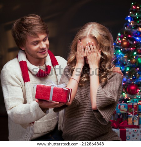 young happy smiling casual couple presenting red gift over christmas tree and lights on background. warm light