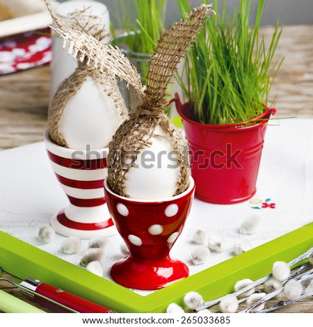 Easter eggs with sackcloth rabbit ears into the red dotted and striped egg cups, pussy willow and germinated grass seeds into the red bucket