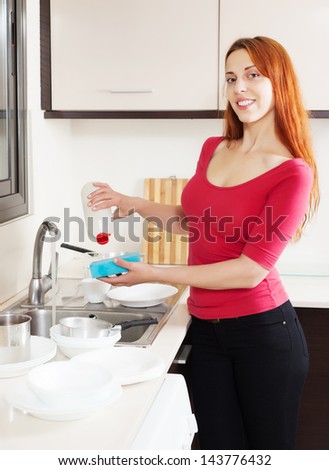 Happy housewife in red washing plates with dishwasher detergent in home kitchen