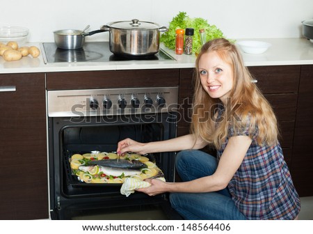 Smiling housewife cooking saltwater fish and potatoes on sheet pan in oven at  kitchen