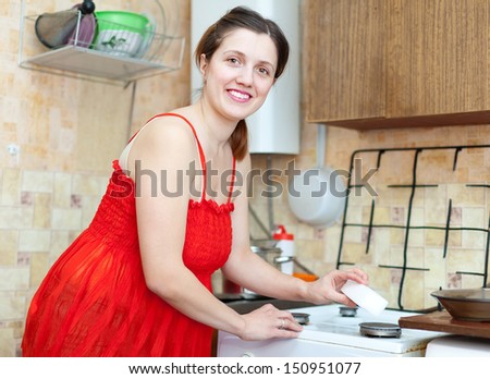 Happy young housewife in red  cleans the gas-stove with melamine sponge  in kitchen at home