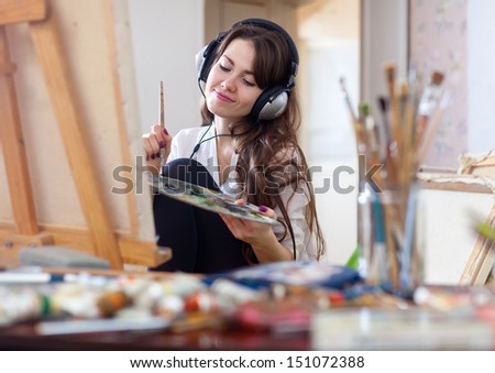 Long-haired woman in headphones  paints with oil colors and brushes on canvas in workshop interior