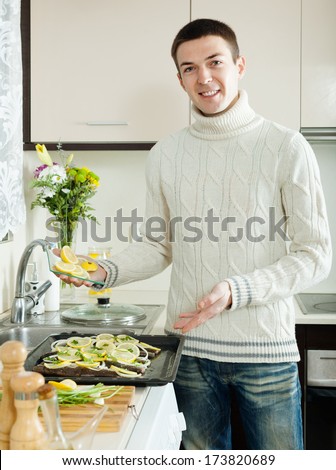 Smiling man putting pieces of lemon in fish at home kitchen