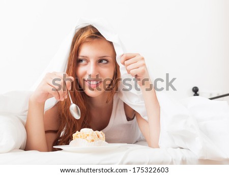 Young woman eating sweet cake under sheet in bed at home