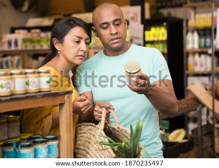 Interested Latin American couple reading product label on jar while choosing groceries in supermarket