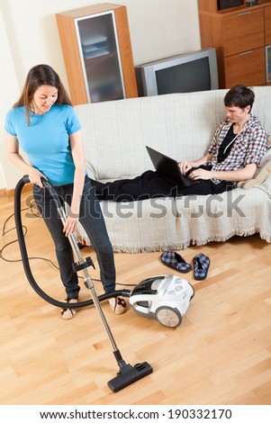man  with laptop  resting over sofa while girl doing house cleaning with vaccuum cleaner