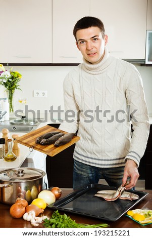 Handsome man putting  raw trout into roasting pan at kitchen table