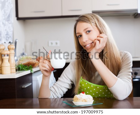 Happy blonde woman drinking tea with cake at table in home kitchen