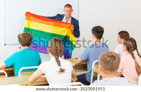 Young progressive teacher discussing with teenage students about LGBT social movements in classroom, holding rainbow flag. Concept of supporting of LGBT or gender identity between youth..