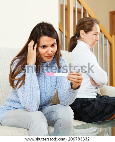 Sad  girl with pregnancy test  against unhappy mature mother at home