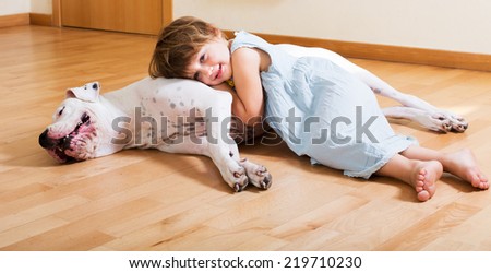 Cheerful cute little girl hugging big white dog lying on the floor at home