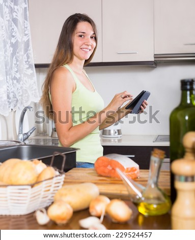 Smiling girl cooking salmon fish and reading   e-book at  kitchen