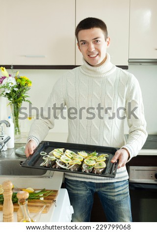 Smiling guy putting pieces lemon in fish at home kitchen