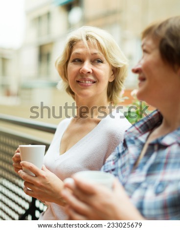 Smiling aged female friends staying on balcony with cup of coffee