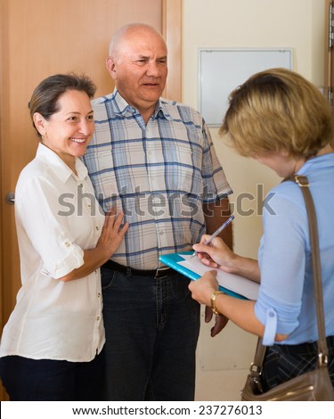 Aged couple answer questions of smiling woman with papers at door in home. Focus on man