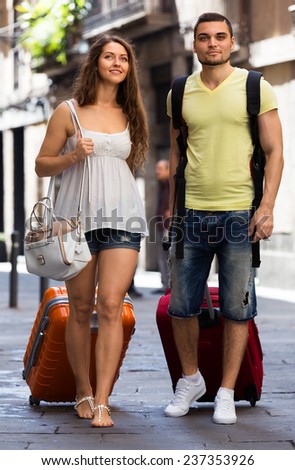 Happy young pair in shorts with luggage walking through city street