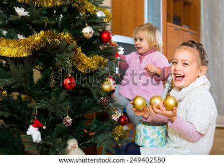Portrait of two happy young girls decorating christmas tree. One of them is holding golden globe ornaments and laughing