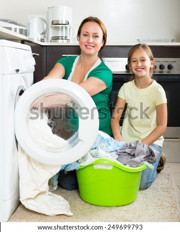 Home family laundry. Happy mother with smiling preschooler daughter loading clothes into washing machine in kitchen