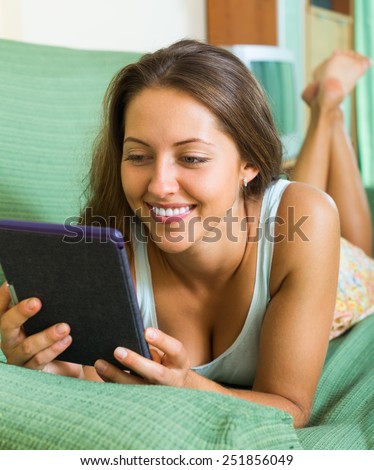 Smiling young woman laying on couch and reading something from tablet screen